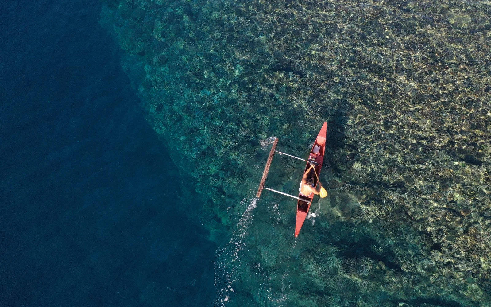 Aerial view of a single outrigger canoe paddling over reef and deep blue water